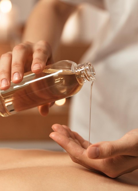Wohltuende Ölmassage Eine Person gießt vorsichtig Massageöl aus einer Glasflasche in ihre Handfläche, während im Vordergrund ein liegender Rücken zu sehen ist. Warme Lichter im Hintergrund schaffen eine entspannte Atmosphäre.