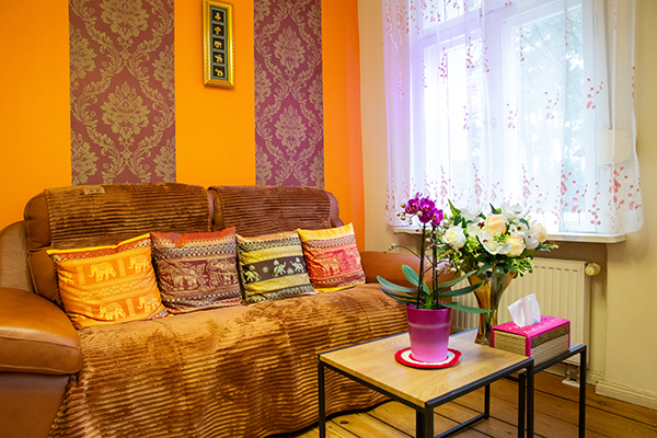 Wartebereich eines Massagesalons mit braunem Sofa, bunten Kissen, floraler Tapete und warmen Orangetönen. Ein Beistelltisch mit Orchidee und ein weiterer Tisch mit Rosenstrauß und Papiertuchbox sorgen für eine einladende Atmosphäre. Ein großes Fenster mit weißen Vorhängen lässt Tageslicht herein.