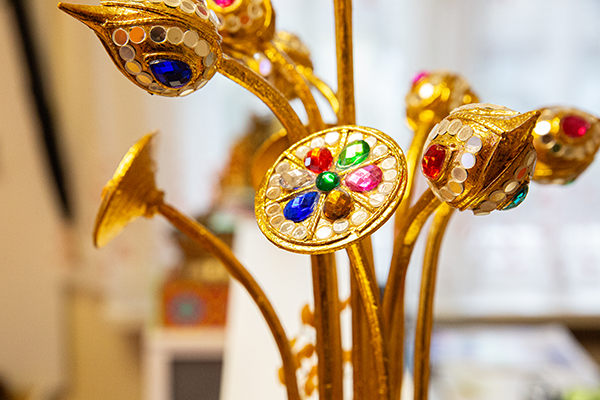 Nahaufnahme einer goldenen Dekoration mit floralen Formen, besetzt mit bunten Schmucksteinen. Im Hintergrund ist ein heller, freundlich gestalteter Raum erkennbar.