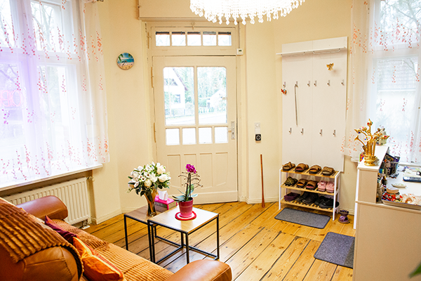 Heller Eingangsbereich eines Massagesalons mit braunem Sofa, kleinen Beistelltischen mit Blumen, einer Garderobe mit Schuhregal und einem funkelnden Kronleuchter. Die Tür mit Glaselementen und dekorierte Fenster lassen viel Tageslicht herein.