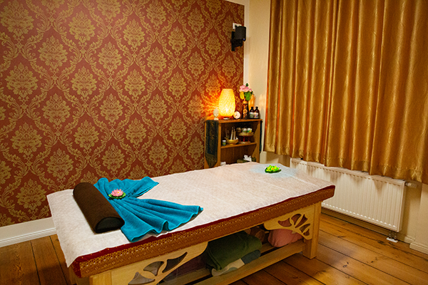 Massageraum mit gemütlicher Massageliege, dekorativer roter Tapete, goldenen Vorhängen und einem Regal mit Massageölen. Eine warme Lichtquelle sorgt für eine entspannte Atmosphäre.