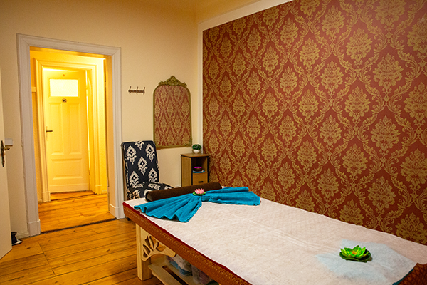 Massageraum mit einer komfortablen Liege, roten floralen Tapeten, einem Spiegel, einem gepolsterten Stuhl und einem kleinen Beistelltisch. Eine geöffnete Tür führt in einen warm beleuchteten Flur.