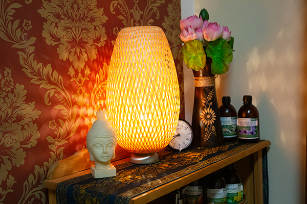 Dekoratives Regal mit leuchtender Tischlampe, Buddha-Statue, Vase mit rosa Lotusblüten und Massageölen. Eine rote florale Tapete bildet den Hintergrund.