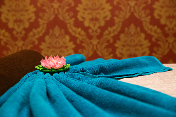 Nahaufnahme einer Massageliege mit blauem Handtuch, rosa Lotusblüte und brauner Nackenrolle, vor einer roten, floral gemusterten Tapete.