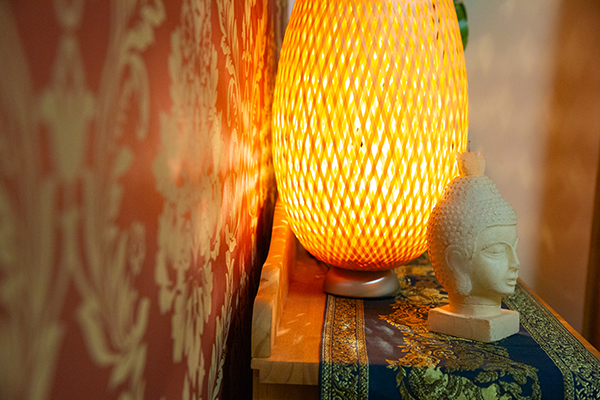 Nahaufnahme einer warm leuchtenden Tischlampe neben einer Buddha-Statue auf einem mit goldenen Ornamenten verzierten Regal, vor einer roten, floralen Tapete.
