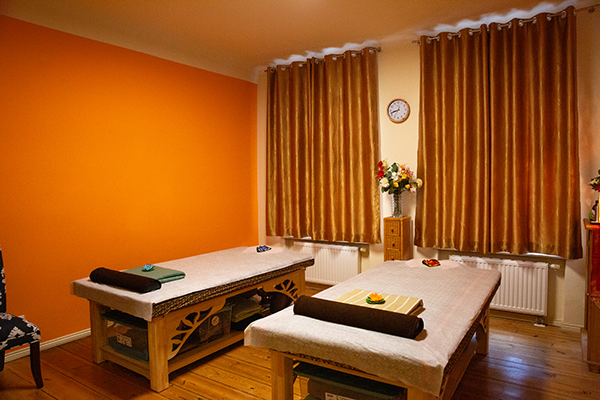 Massageraum mit zwei Liegen, braunen Nackenrollen, dekorativen Blumen, einer orangen Wand, goldenen Vorhängen und einer Holzkommode mit Blumenvase. Eine ruhige und entspannende Atmosphäre.