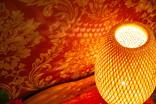 Nahaufnahme einer warm leuchtenden Tischlampe mit geflochtener Struktur, die ein Schattenspiel auf eine rote, floral gemusterte Tapete wirft.