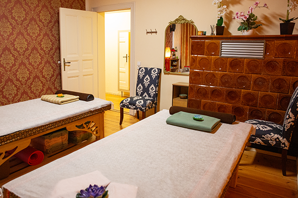 Massageraum mit zwei Liegen, roten floralen Tapeten, traditionellem Kachelofen, Spiegel, Kommode mit Massageutensilien und gepolsterten Stühlen. Eine geöffnete Tür führt in einen warm beleuchteten Flur.