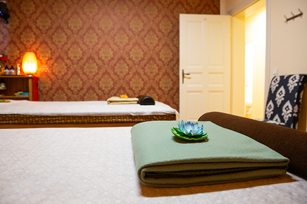 Massageraum mit zwei Liegen, roten floralen Tapeten, einer gefalteten grünen Decke mit Lotusblüte, brauner Nackenrolle und warm beleuchtetem Flur im Hintergrund.