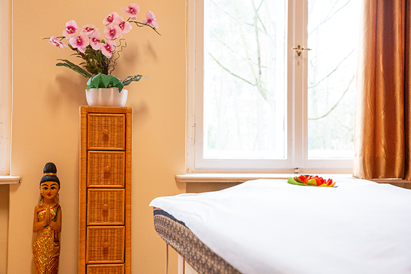 Massageliege mit weißer Auflage, dekorativer Lotusblüte, Rattan-Kommode mit Orchideenpflanze und goldfarbener Holzfigur vor einem großen Fenster mit natürlichem Tageslicht.