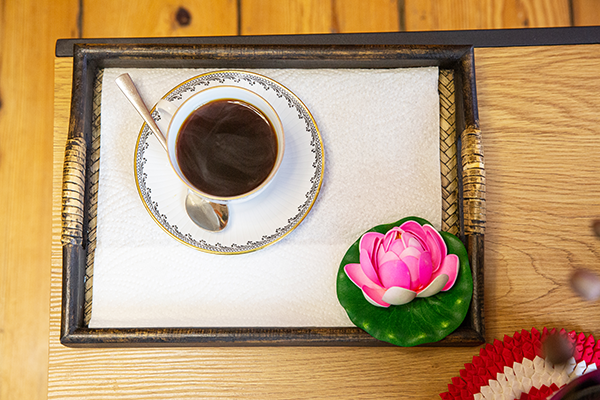 Tablett mit einer Tasse Kaffee oder Tee in einer weißen Porzellantasse mit goldener Verzierung, einem kleinen Löffel und einer rosa Lotusblüte, auf einem Holztisch arrangiert.